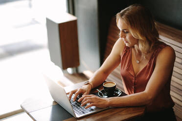 Freelancer sitting at a coffee table working on laptop with a cup of coffee on the table. Woman doing her work sitting at a coffee shop. - JLPSF13783