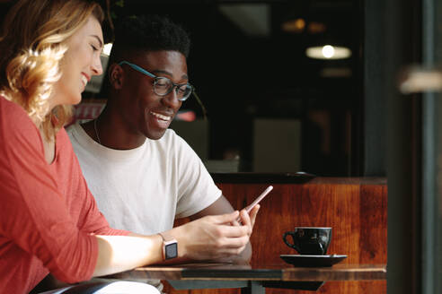 Mann und Frau sitzen an einem Kaffeetisch und schauen auf ein Mobiltelefon. Glückliches Paar in einem Café, das ein Mobiltelefon mit einer Tasse Kaffee auf dem Tisch benutzt. - JLPSF13786