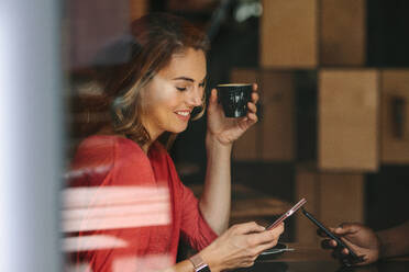 Frau schaut auf ihr Handy, während sie eine Kaffeetasse hält. Lächelnde Frau, die sich mit ihrer Freundin in einem Café trifft. - JLPSF13788