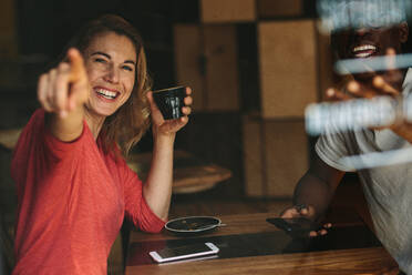 Lächelnde Frau, die auf etwas zeigt, das an einem Kaffeetisch sitzt und eine Kaffeetasse hält. Lächelnde Frau, die mit ihrem Freund in einem Kaffeehaus sitzt. - JLPSF13790