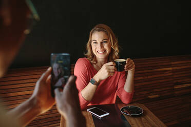 Woman sitting at a coffee table posing for a photograph holding a coffee cup. Man taking photo of a woman using mobile phone at a coffee shop. - JLPSF13792