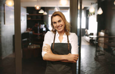 Happy coffee shop owner standing at the entrance of her shop. Female barista standing at a café wearing apron and arms crossed. - JLPSF13803