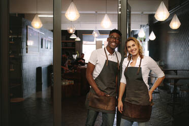 Ein glückliches Café-Besitzerpaar steht in seinem Laden. Ein Mann und eine Frau stehen in ihrem Café und tragen eine Schürze. - JLPSF13805