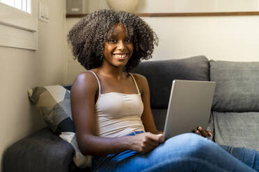 Glückliche Frau mit Laptop auf dem Sofa im Wohnzimmer sitzend - DLTSF03247
