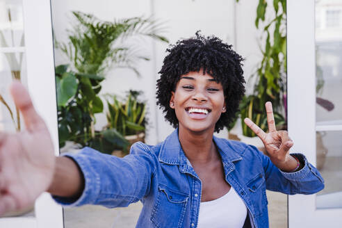Glückliche Frau zeigt Friedenszeichen und macht ein Selfie in der Tür - EBBF06588