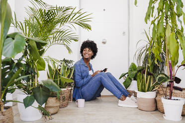 Glückliche junge Frau mit Smartphone inmitten von Pflanzen sitzend - EBBF06596