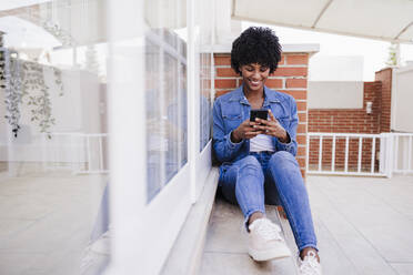 Glückliche Frau mit Smartphone auf Veranda sitzend - EBBF06604