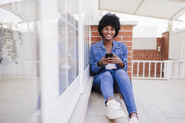 Happy young woman with smart phone sitting on porch - EBBF06605