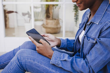 Smiling woman using smart phone sitting by door - EBBF06606