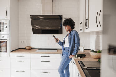 Glückliche Frau, die ein Smartphone benutzt und sich zu Hause auf die Küchentheke stützt - EBBF06609