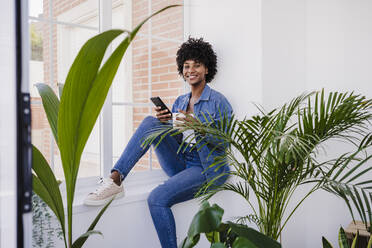 Happy woman with smart phone sitting on window sill by plants at home - EBBF06612