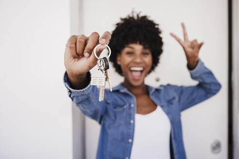 Fröhliche Frau zeigt Hausschlüssel und Friedenszeichen-Geste vor der Tür - EBBF06620