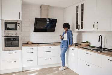 Young woman using smart phone leaning on kitchen counter at home - EBBF06635