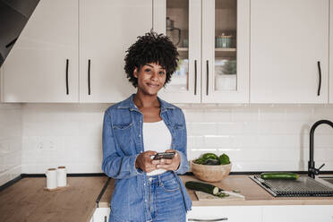 Lächelnde junge Frau mit Smartphone, die sich zu Hause an die Küchentheke lehnt - EBBF06637