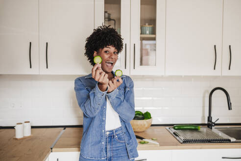 Fröhliche junge Frau zeigt Zucchinischeiben, die auf dem Küchentisch zu Hause liegen - EBBF06641