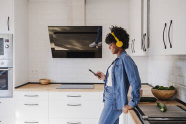 Smiling young woman wearing wireless headphones standing by kitchen counter at home - EBBF06663