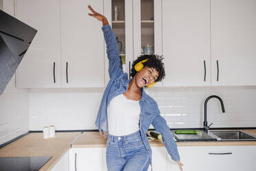 Cheerful young woman wearing wireless headphones dancing in kitchen at home - EBBF06667