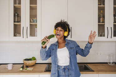 Cheerful woman wearing wireless headphones singing with zucchini in kitchen at home - EBBF06668