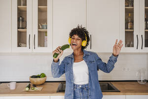 Fröhliche Frau mit drahtlosen Kopfhörern singt mit Zucchini in der Küche zu Hause - EBBF06668