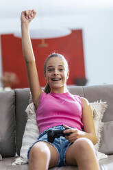Fröhliches Mädchen mit erhobenem Arm sitzt mit Joystick auf dem Sofa zu Hause - JSRF02228