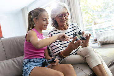 Granddaughter talking to grandmother holding joystick on sofa at home - JSRF02232