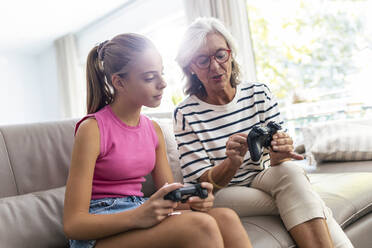 Grandmother showing game controller to granddaughter sitting on sofa at home - JSRF02233