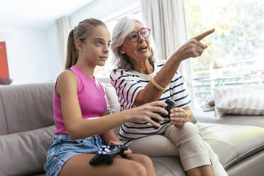 Grandmother pointing by granddaughter sitting with joystick on sofa at home - JSRF02235