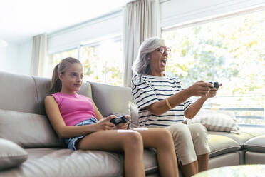 Excited grandmother playing video game with granddaughter sitting on sofa at home - JSRF02240