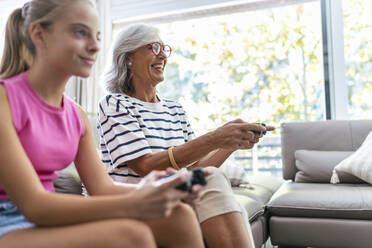 Happy grandmother playing video game with granddaughter at home - JSRF02242