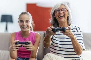 Happy grandmother and granddaughter playing video game with joysticks at home - JSRF02245