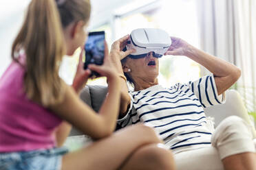 Girl photographing grandmother wearing virtual reality headset at home - JSRF02246
