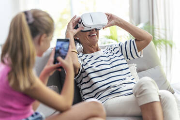 Enkelin fotografiert ältere Frau, die zu Hause ein Virtual-Reality-Headset benutzt - JSRF02247