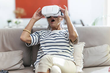 Senior woman with mouth open wearing virtual reality headset sitting on sofa at home - JSRF02248