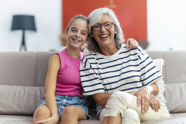Glückliches Mädchen sitzt mit Arm um Großmutter auf Sofa zu Hause - JSRF02250