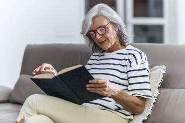 Senior woman wearing eyeglasses reading book at home - JSRF02251