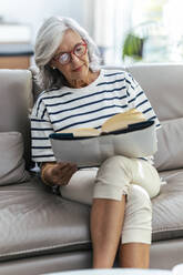 Frau, die mit gekreuzten Beinen auf dem Sofa zu Hause sitzt und ein Buch liest - JSRF02253