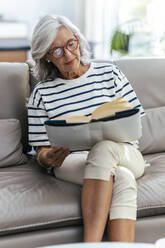 Woman reading book sitting with legs crossed on sofa at home - JSRF02253