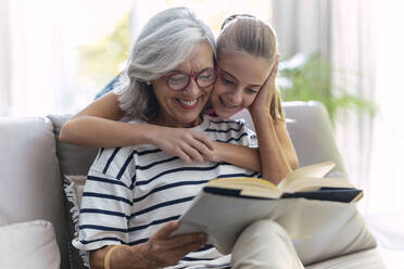 Lächelndes Mädchen umarmt Großmutter beim Lesen eines Buches auf dem Sofa zu Hause - JSRF02254