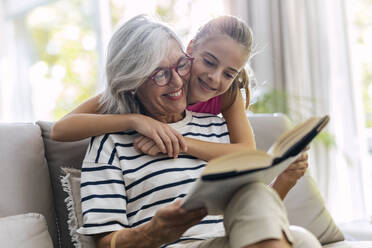 Grandmother reading book with granddaughter on sofa at home - JSRF02255