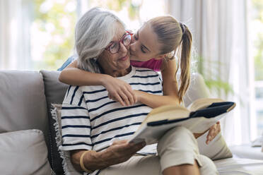 Girl kissing grandmother reading book sitting on sofa at home - JSRF02256