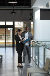 Smiling colleagues discussing over document on clipboard in office corridor - AMNF00023