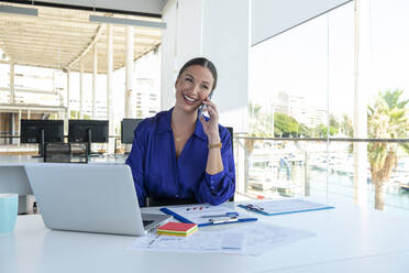 Lächelnde Geschäftsfrau mit Laptop im Gespräch auf dem Smartphone am Schreibtisch im Büro - AMNF00035