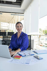 Smiling businesswoman with laptop on desk at work place - AMNF00036