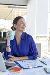 Lächelnde junge Geschäftsfrau mit Kaffeetasse am Schreibtisch im Büro - AMNF00037
