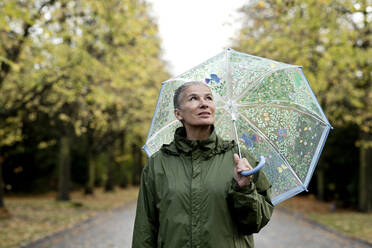 Nachdenkliche ältere Frau mit Regenmantel und Regenschirm - FLLF00805