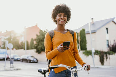 Lächelnde Frau mit lockigem Haar, die ein Mobiltelefon und ein Fahrrad auf der Straße hält - EBBF06686