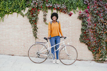 Smiling woman with bicycle in front of ivy wall - EBBF06689