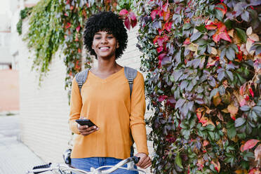 Lächelnde junge Frau mit Smartphone bei Pflanzen an der Wand - EBBF06690