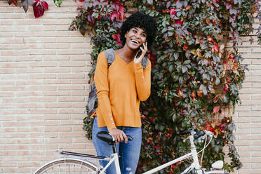 Glückliche Frau mit Fahrrad, die vor einer Efeumauer mit ihrem Smartphone telefoniert - EBBF06692