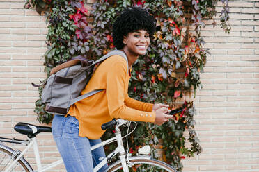 Smiling young woman with backpack holding smart phone on bicycle - EBBF06693